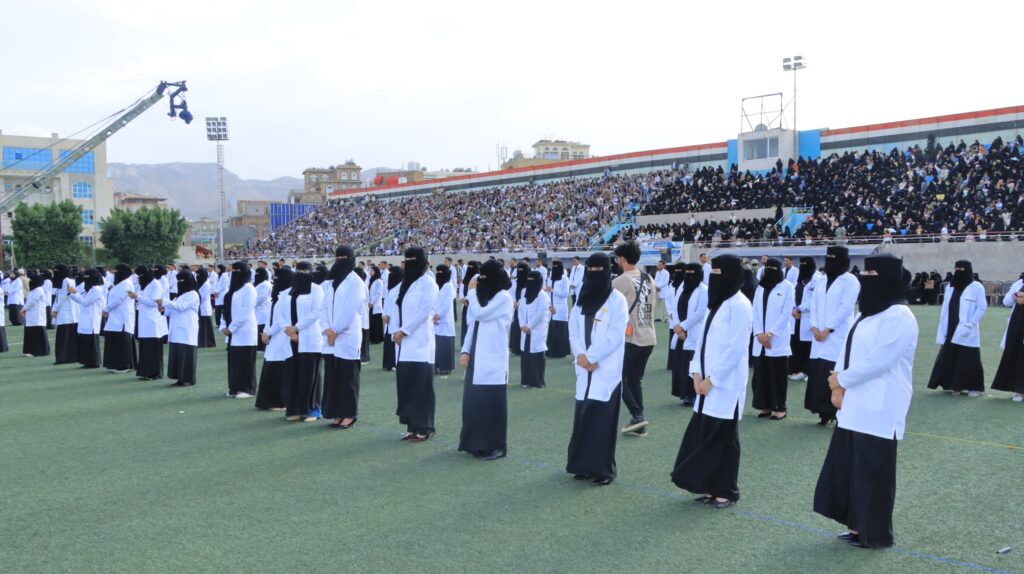 1003984333 - جامعة 21 سبتمبر Large group of women in white lab coats and black hijabs lined up on a sports field during a ceremony, with a packed stadium in the background.