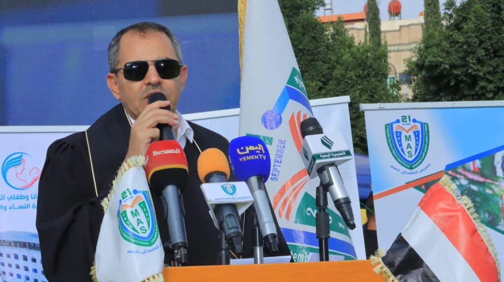 1003984338 - جامعة 21 سبتمبر Religious figure wearing sunglasses speaks into multiple microphones at an outdoor press conference with banners and flags in the background.