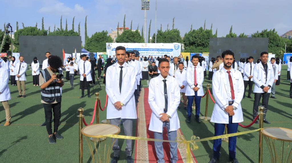 1003984346 - جامعة 21 سبتمبر Group of people in white lab coats standing on a sports field during an outdoor ceremony, with a red carpet, drums, and photographers in the foreground.