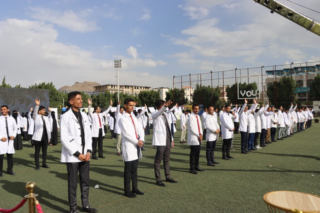 1003984347 - جامعة 21 سبتمبر Group of people in white lab coats standing in rows on a sports field during a ceremony, with raised arms and a blue sky above.