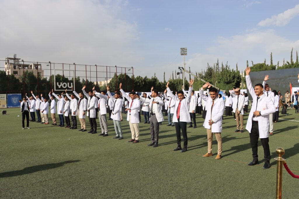 1003984348 - جامعة 21 سبتمبر Group of people in white lab coats standing in a line on a sports field, waving at the camera.