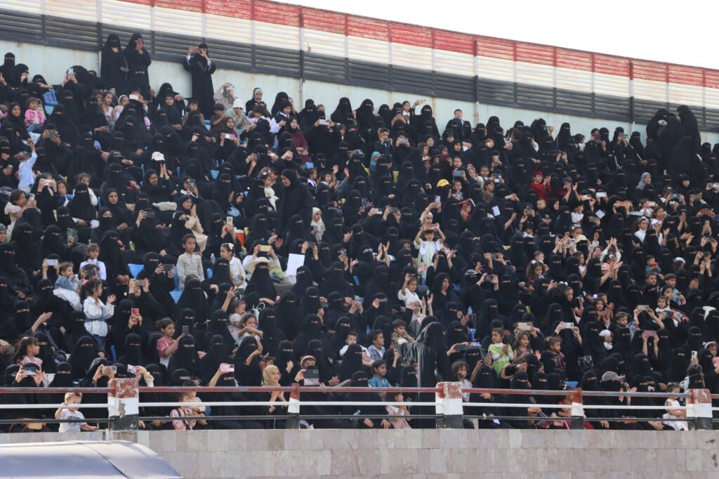 1003984349 - جامعة 21 سبتمبر Crowd of women in black niqab covering faces, seated in stadium stands, many holding up smartphones to take photos or video, with some children visible throughout.