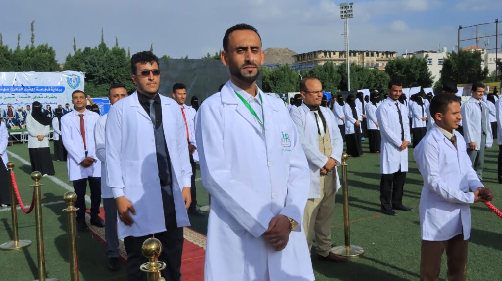 1003984350 - جامعة 21 سبتمبر Group of men in white lab coats at an outdoor ceremony on a field, with banners and a crowd in the background.