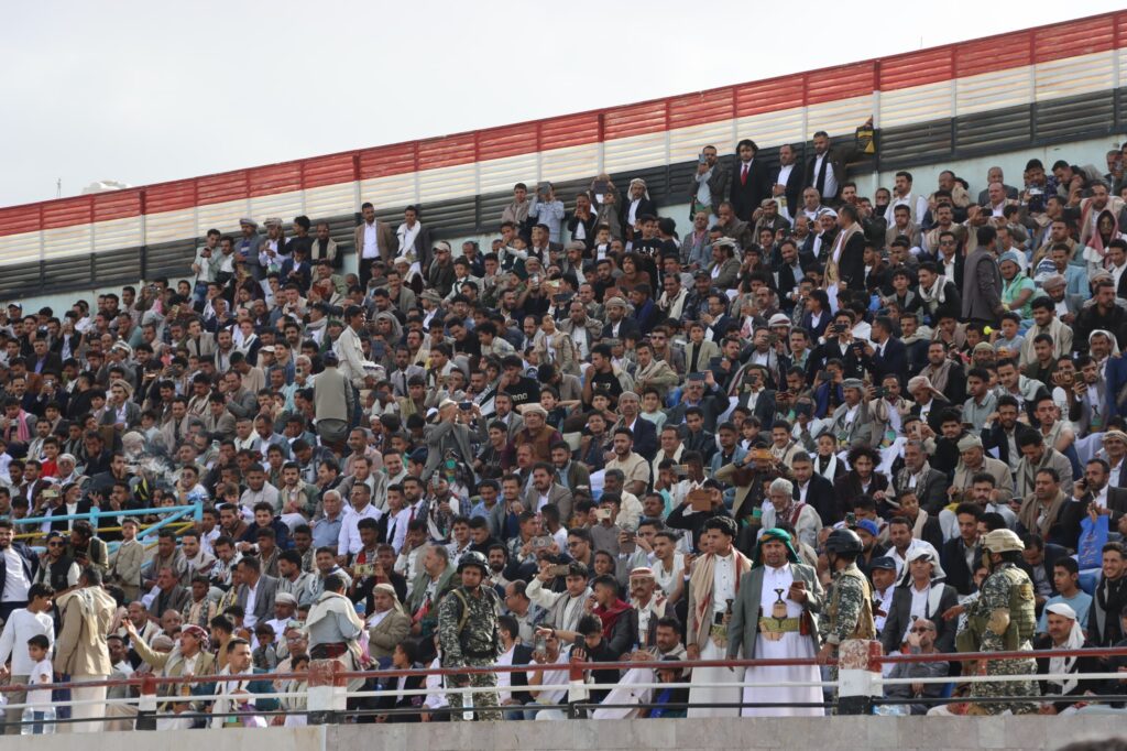 1003984351 - جامعة 21 سبتمبر Large crowd of spectators seated in stadium stands, many holding up phones to record the event.