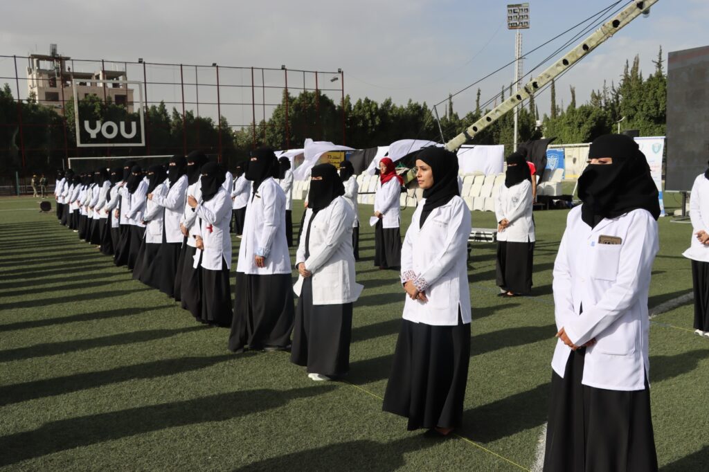 1003984352 - جامعة 21 سبتمبر Row of women in white lab coats and black hijabs standing in formation on a sports field during an outdoor ceremony.