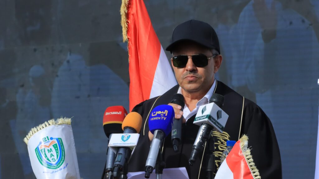 1003984482 - جامعة 21 سبتمبر Man wearing a black cap and sunglasses speaks into several microphones at a press conference, with Yemeni flags in the background.