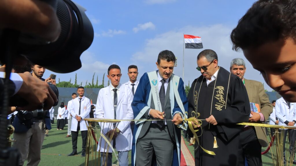 1003984483 - جامعة 21 سبتمبر Group of graduates in gowns cutting a ceremonial ribbon outdoors during a graduation event with a flag in the background