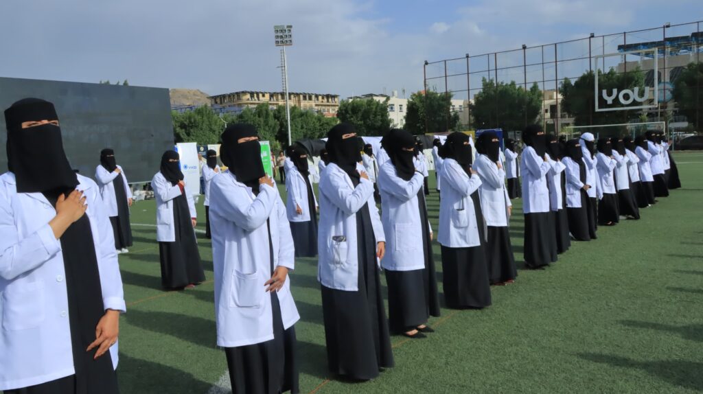 1003984484 - جامعة 21 سبتمبر Group of women in white lab coats and black niqabs standing in neat rows on a sports field, hands over their hearts.
