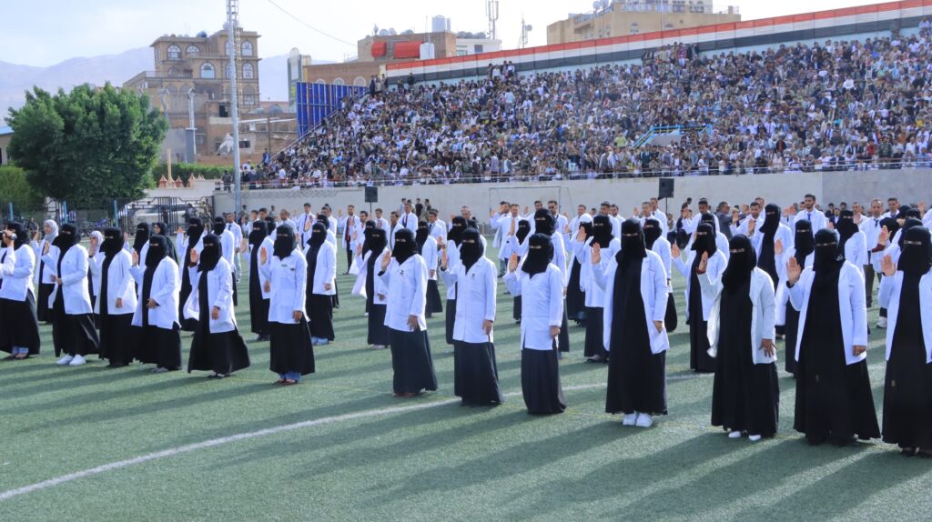 1003984485 - جامعة 21 سبتمبر Large crowd watches a parade of students in white coats and black hijabs marching on a sports field, with stands full of spectators behind them.