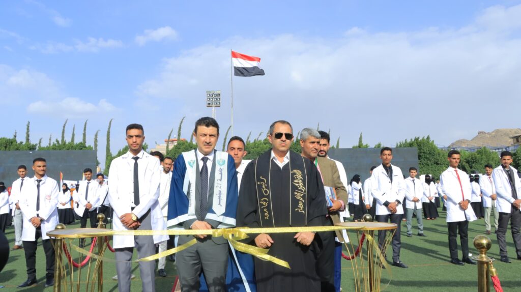 1003984486 - جامعة 21 سبتمبر Graduates in white coats gather on a field for an outdoor ceremony, with an Egyptian flag rising behind them.