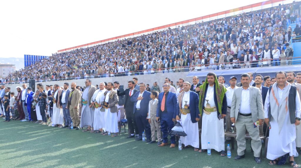 1003984487 - جامعة 21 سبتمبر Large crowd of spectators in a stadium, standing on the field and in the bleachers during an event, many in traditional clothing and suits
