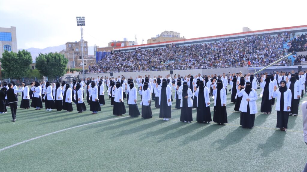 1003984490 - جامعة 21 سبتمبر Crowd in stadium stands watches a field ceremony with students in white coats and black hijabs lined up on the field.