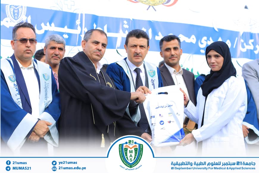 1003985631 - جامعة 21 سبتمبر Group of men in robes and a woman in a white lab coat exchange a certificate at a university ceremony, with a blue banner and emblem in the background.