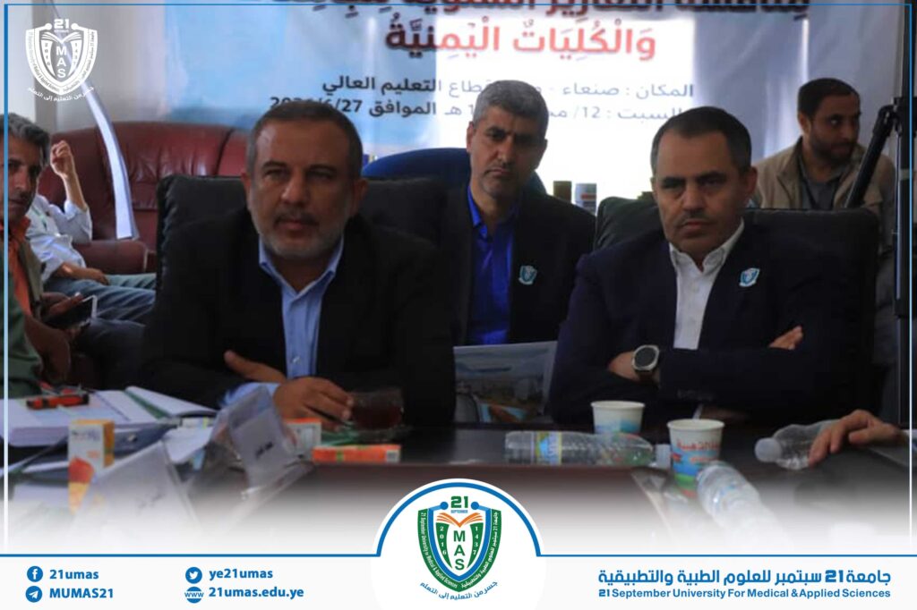 Several men in suits sit around a conference table during a meeting, with a blue banner and a university crest in the background.