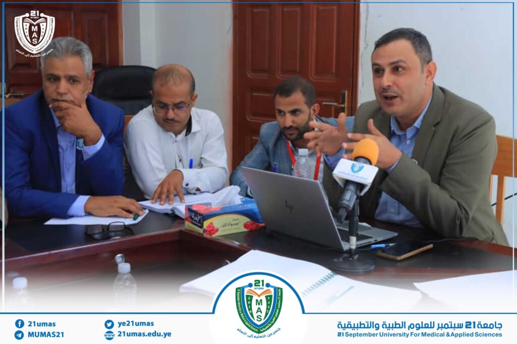 Panel of four men in a meeting at a university event, with a laptop and microphone on the table and a logo in the foreground reading 21 September University for Medical & Applied Sciences, Yemen (MUMAS).