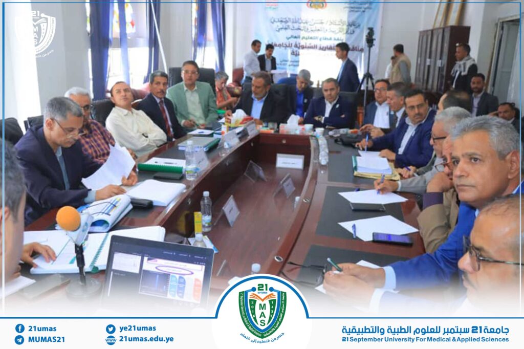 Group of professionals seated around a long conference table in a university meeting room, with papers, notebooks, and microphones; a blue banner and university logo visible in the background.