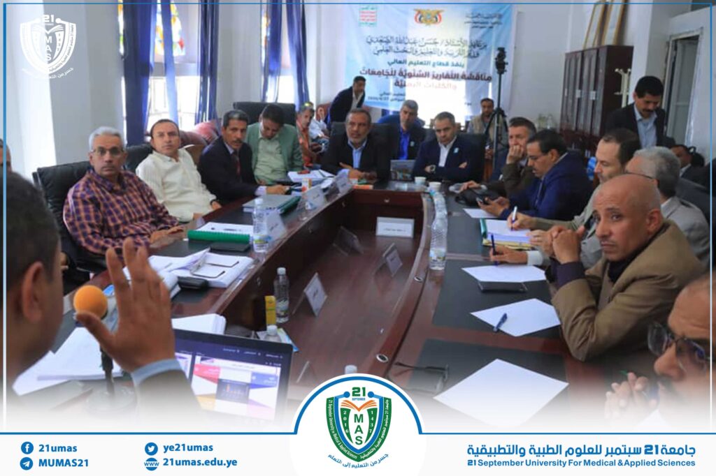 Group of professionals seated around a conference table in a meeting room with a banner in Arabic in the background, at 21 September University for Medical & Applied Sciences event.