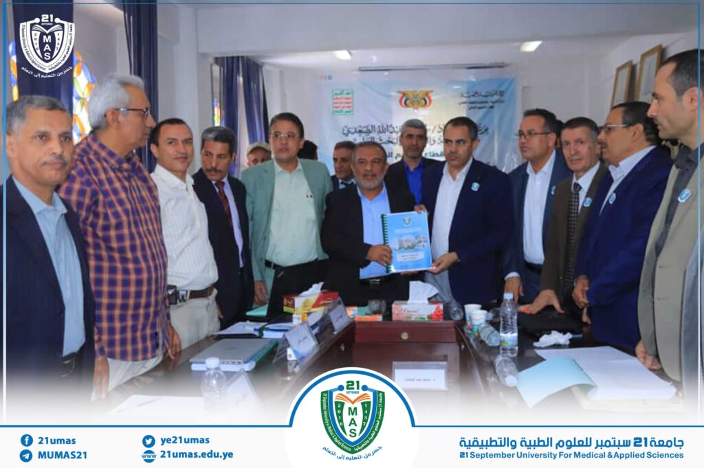 Group of men in suits and shirts gathered around a conference table, some holding a blue booklet, with a banner and university logo in the background.