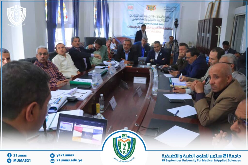 Group of professionals around a large conference table in a meeting room, with a blue event banner in the background and university logos visible.
