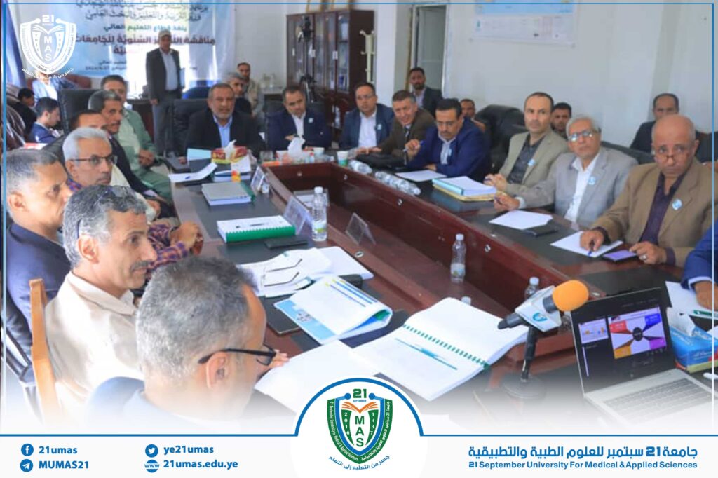 A group of men seated around a large conference table in a university meeting room, with university logos visible on the wall/codium display in the background.