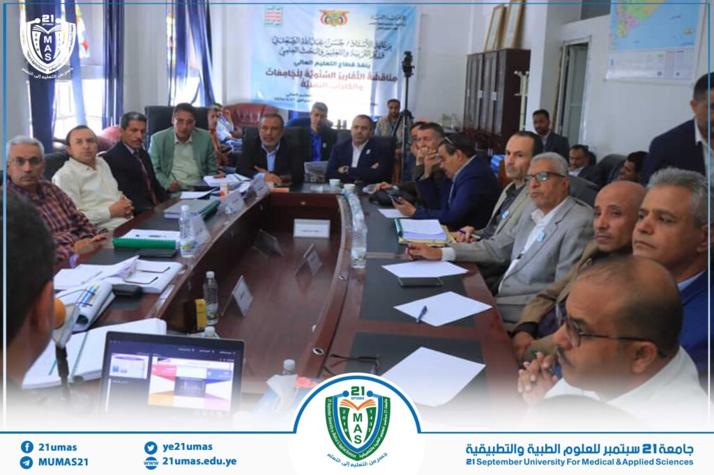 Group of officials seated around a long conference table in a meeting room, with banners and university logos in the background.