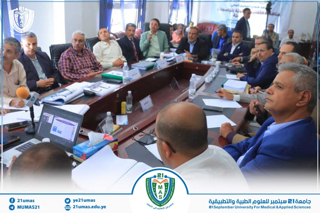 People seated around a long conference table in a meeting room, with laptops, papers, and water bottles; university emblem in the background.