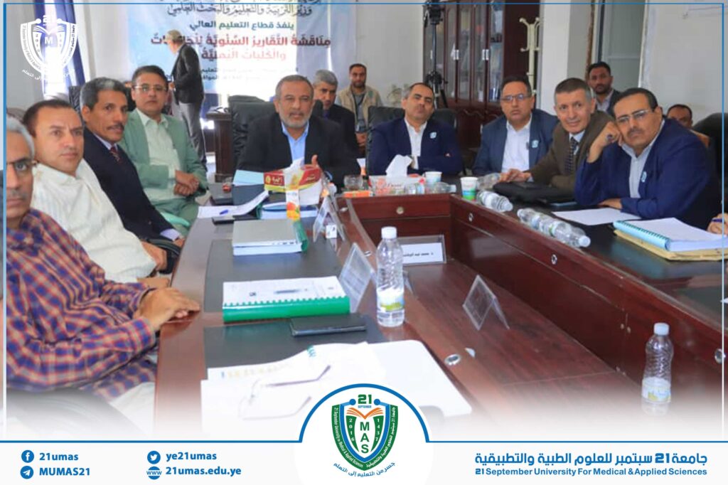 Group of men in formal attire sit around a conference table during a university meeting, with a banner in Arabic behind them and a circular campus logo visible at the bottom center.