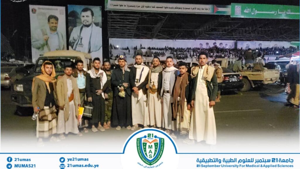 Group of men in traditional Yemeni clothing posing for a night-time group photo at an outdoor event with banners and cars behind.