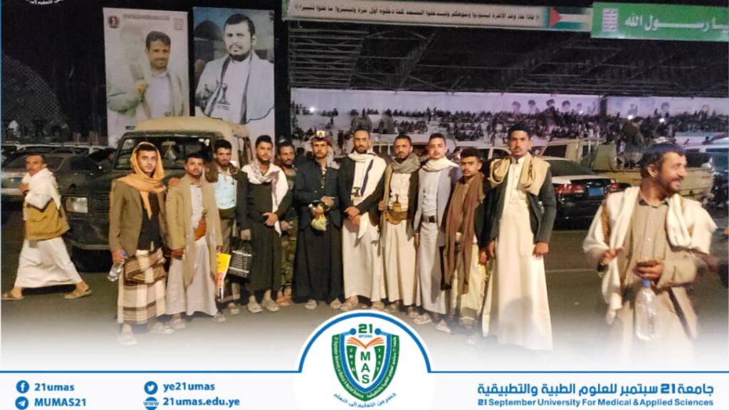 Group of Yemeni men in traditional clothing posing together outdoors at night, with vehicles and large banners in the background and a university logo at the bottom center.