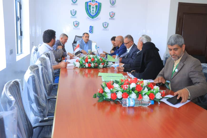People seated around a long conference table in a formal meeting; centerpiece of red and white flowers, papers, and water bottles on the table.