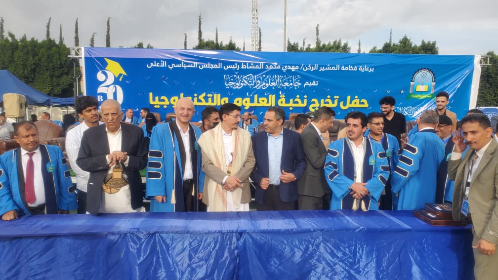 Group of men in blue ceremonial robes at a graduation ceremony in front of a blue banner with Arabic text Students and officials on stage at a graduation event with a long blue table and banner behind them Ceremony scene: graduates in blue gowns standing with officials under a large blue banner African/Arab men in formal attire and blue robes posing on a stage for a graduation ceremony Event photo of graduates and dignitaries at a university commencement, with a blue backdrop banner