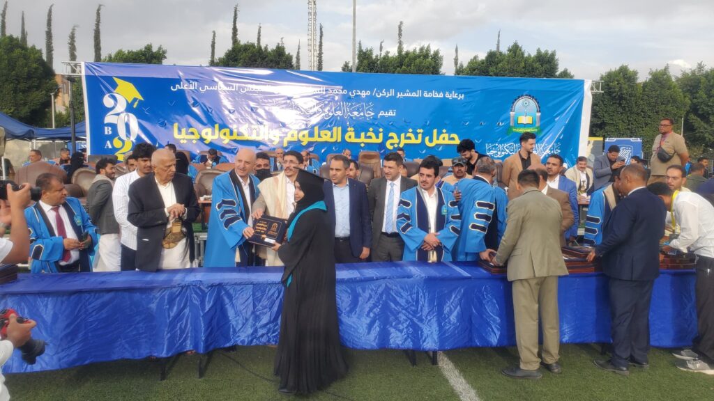 Outdoor graduation ceremony with graduates in blue robes and sashes, presenting diplomas under a large blue banner with Arabic text.
