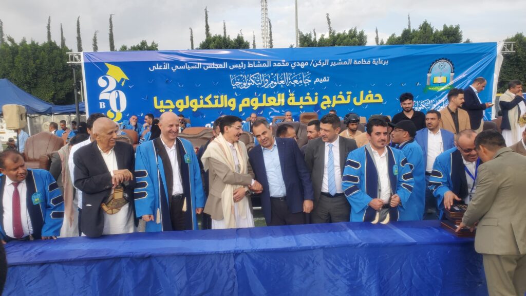 Group of men in blue graduation gowns standing and conversing at an outdoor ceremony with a large blue banner backdrop.