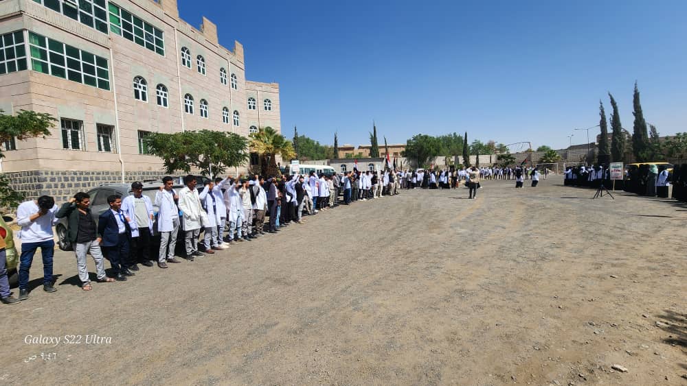 Long line of students in white coats standing along a gravel lot outside a large school building on a sunny day.