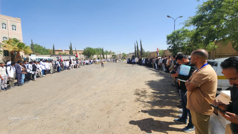 Long line of people standing along a dusty street on both sides during an outdoor ceremony, with buildings and trees in the background.