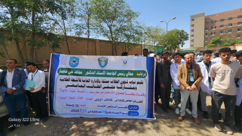 Group of people standing outdoors holding a large blue banner with Arabic text, flanked by trees and a building in the background.