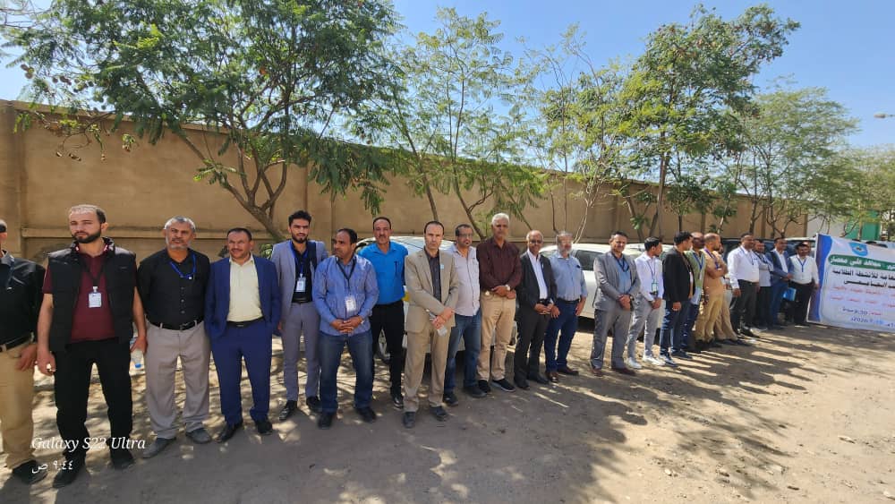 Group of men in business casual attire standing in a line outdoors beside a banner for an event.