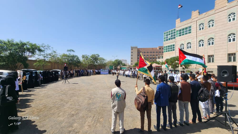 Students gather in a school courtyard for a flag ceremony, with flags held by a line of participants and a speaker at a podium.