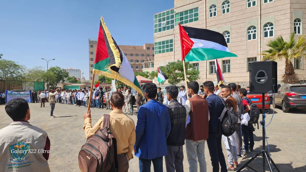 Group of students lined up outdoors at a campus event, holding Palestinian and other flags, with a speaker addressing them and a building in the background.