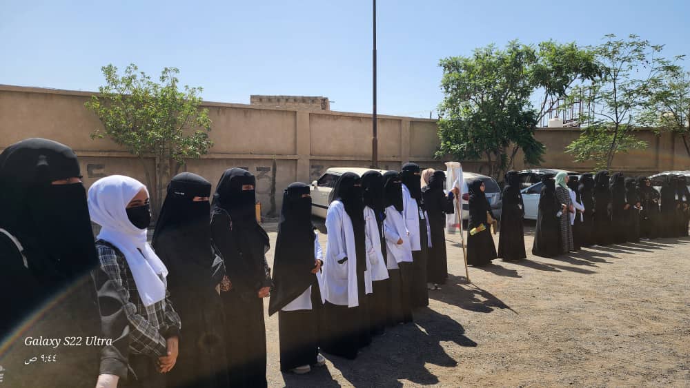 Line of women wearing black abayas and niqabs, some in white coats, standing in a sunlit outdoor area near a wall and parked cars.