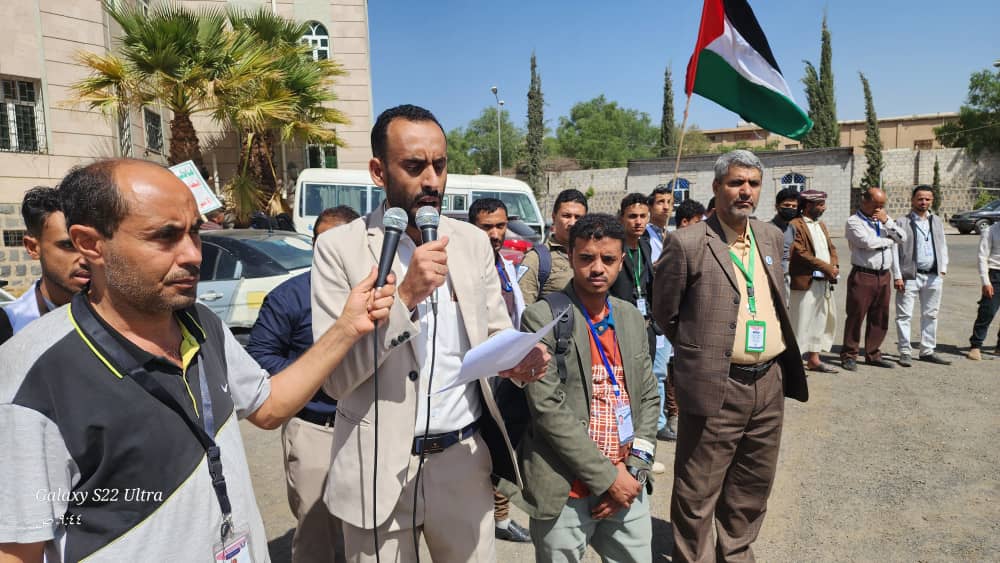 Man in a beige suit speaks into two microphones at an outdoor gathering, with a Palestinian flag visible in the background and people listening behind him.