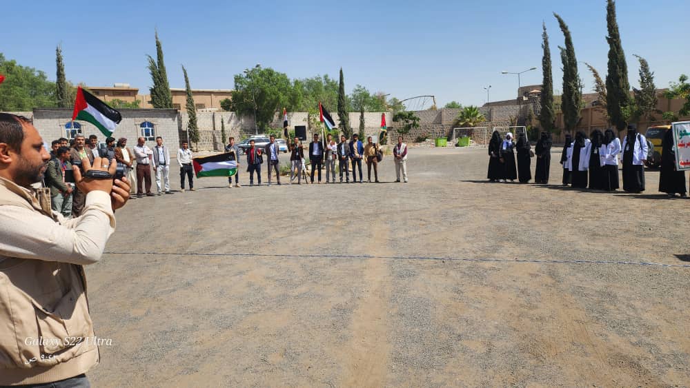 Group of people lined up outdoors, holding Palestinian flags while a man films the scene with a camera on the left.