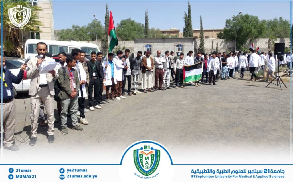 Line of students and faculty standing in a row at an outdoor university event, with a speaker at left and a Palestinian flag, buildings in the background.