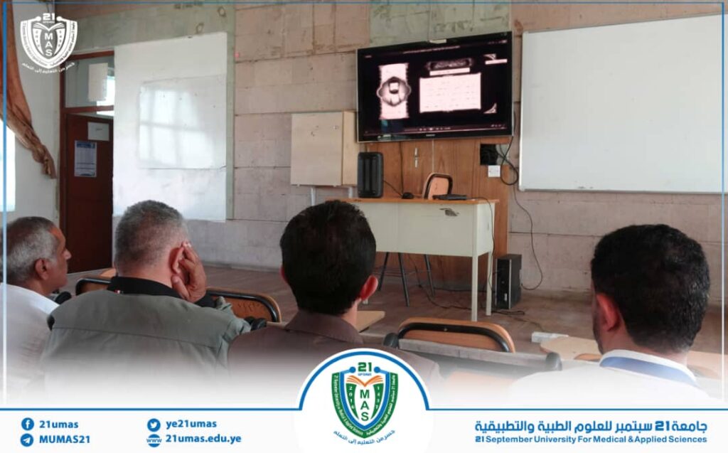 Lecture hall with several men seated, watching a presentation on a wall-mounted screen; university logo overlay at bottom.