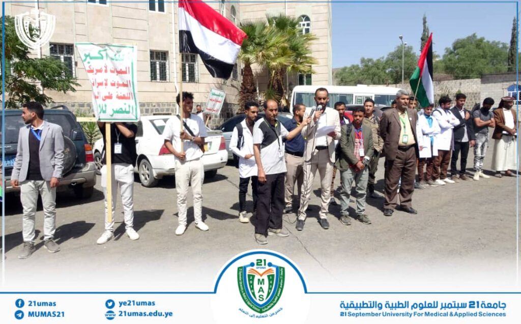 Group of men outdoors, some holding Arabic protest signs and a flag, a man speaks into a microphone.