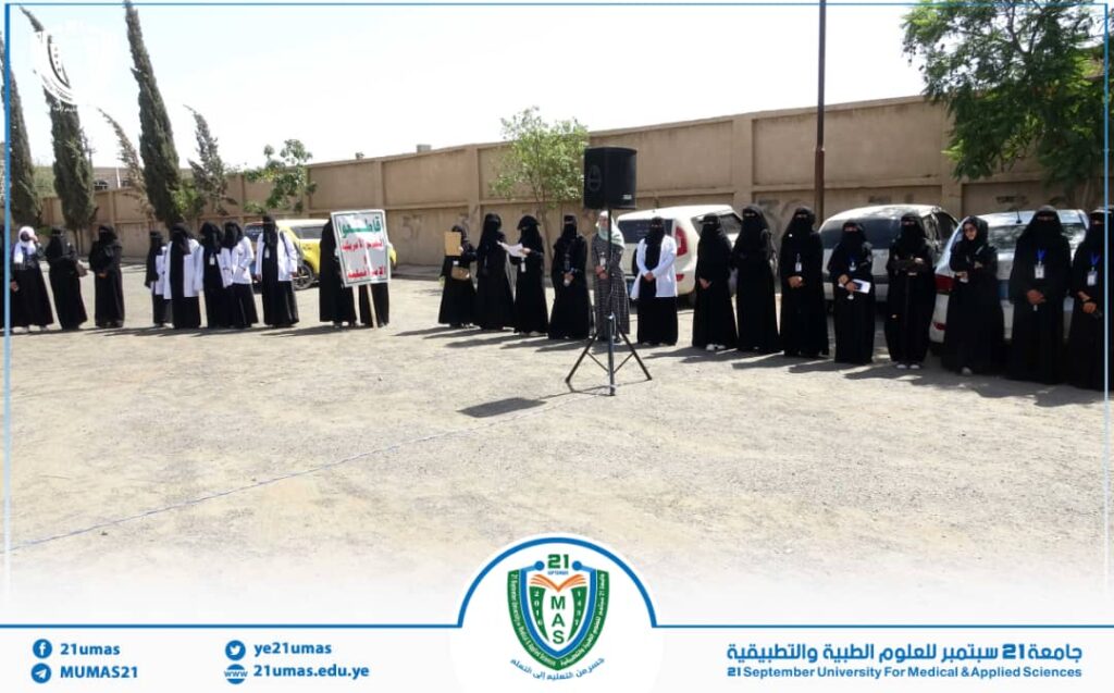Line of women in black abayas standing outdoors for a university event, with a speaker on a stand and a banner in the background.