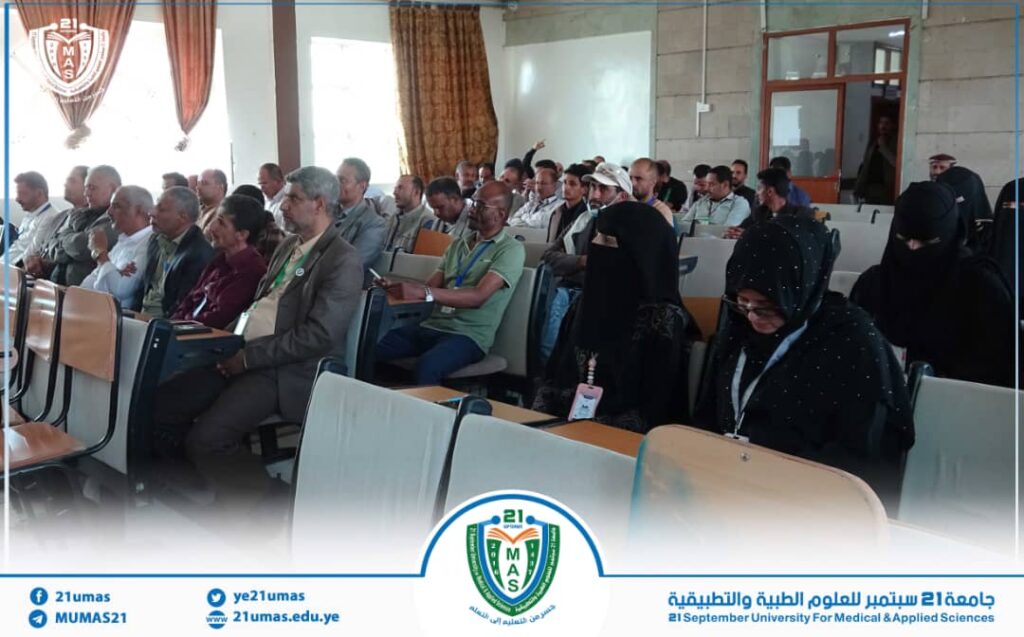 Audience seated in a university lecture hall listening to a presentation at 21umas, with a circular logo banner in the foreground.