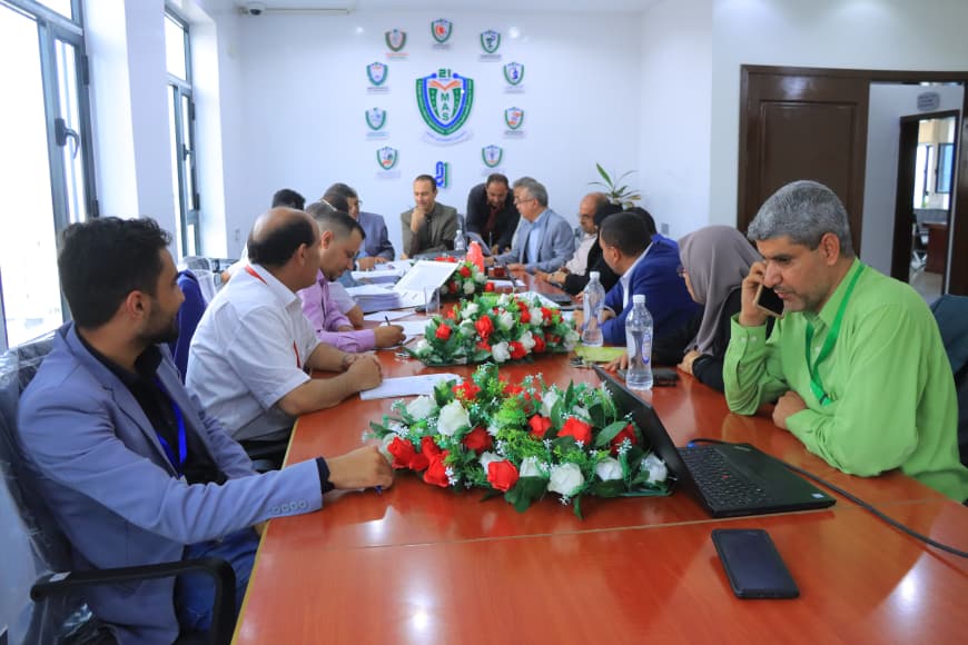 Groups of professionals around a conference table discussing documents, with a floral centerpiece and crest logos on the wall.
