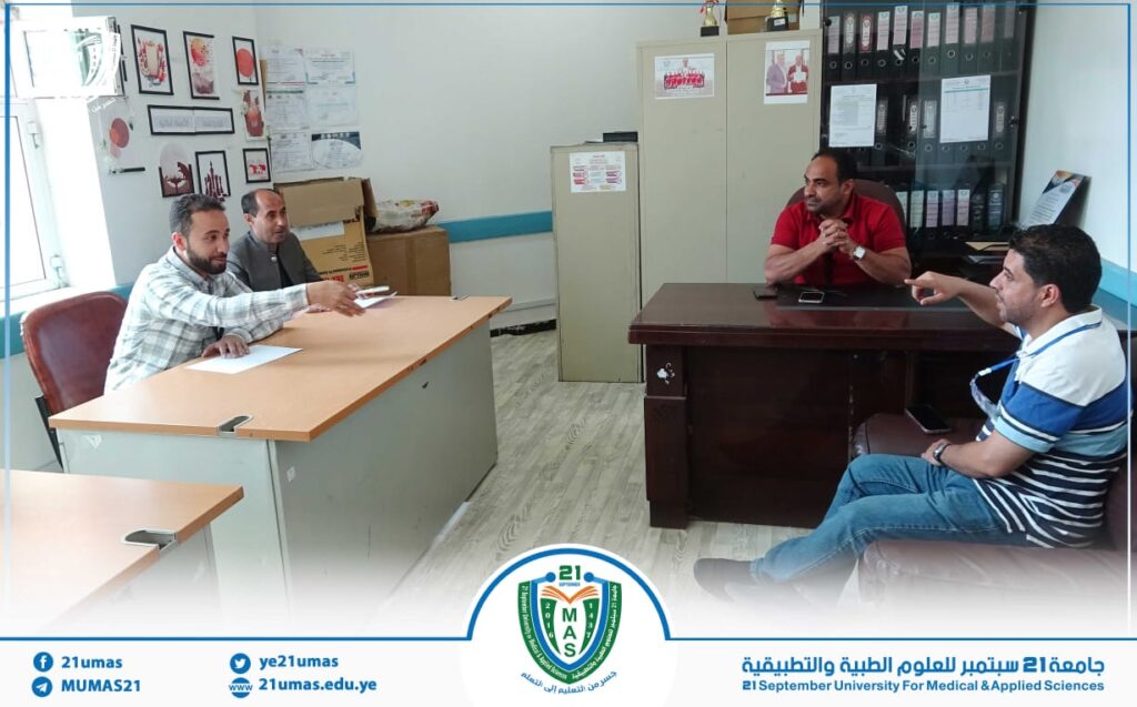 Four men in a casual meeting in an office, with two at a light wood desk on the left and two at a dark desk on the right; university banner at the bottom.Let's provide more concise options.