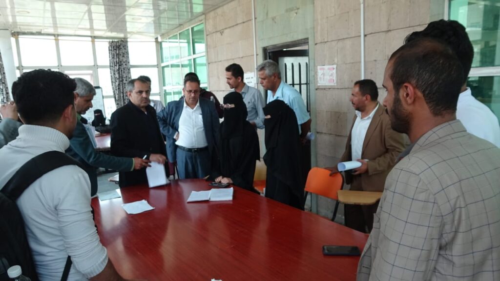 Group of people around a red conference table, examining documents in a bright meeting room, some standing and some seated.